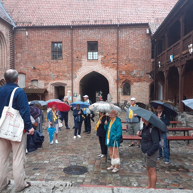 Na dziedzińcu zamku Krzyżackiego widać stare i odrestaurowane mury oraz bramę wjazdową. Beneficjenci słuchają uważnie co opowiada im przewodnik. Mają parasole, które chronią ich od lekkiego deszczu.