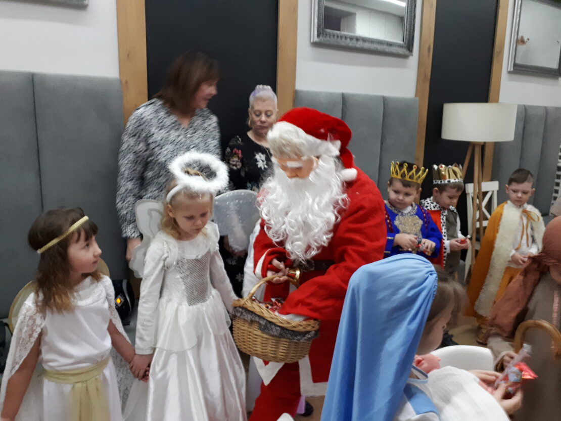 Na środku zdjęcia jest święty Mikołaj. Trzyma lewą rękę w koszyku, czegoś tam szuka. Z jego prawej strony stoją dziewczynki przebrane za aniołki. Mikołaj jest zwrócony w ich stronę. Za nim stoja chłopcy przebrani za Króli.
