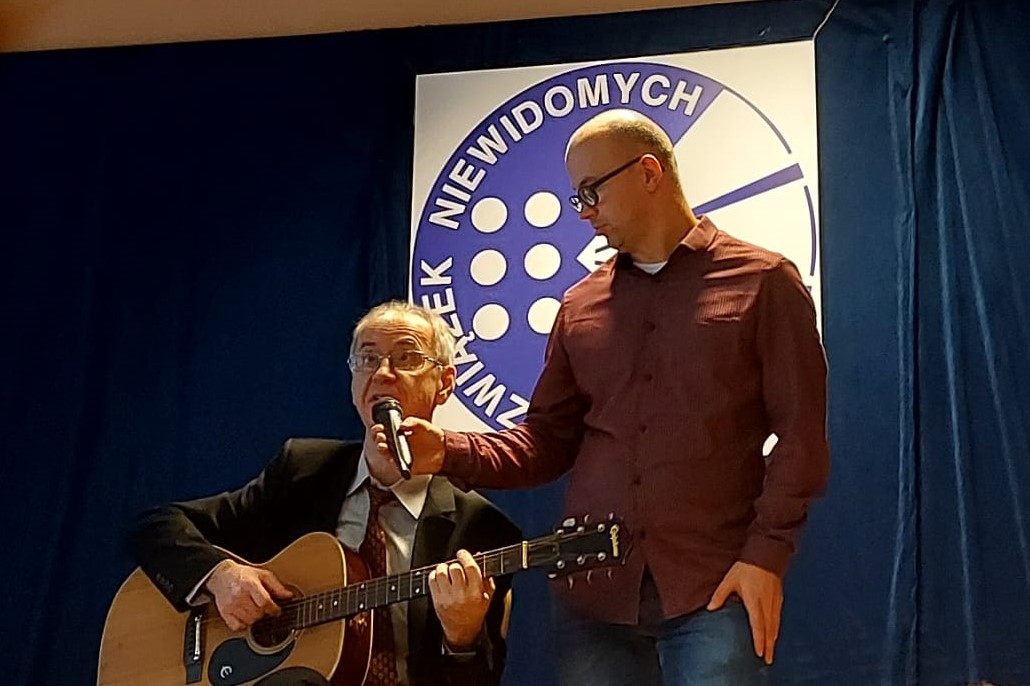 Na scenie siedzi Pan z Ukrainy gra na gitarze  i śpiewa. Pan w bordowej koszuli trzyma mikrofon przed twarzą muzyka.