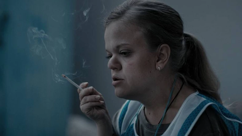 Widok na kobiete paląca papierosa. Ma lekko przymróżone oczy, nad czyms myśli. Szare tło do okoła w prawej ręce trzyma papierosa z którego unosi si dym.