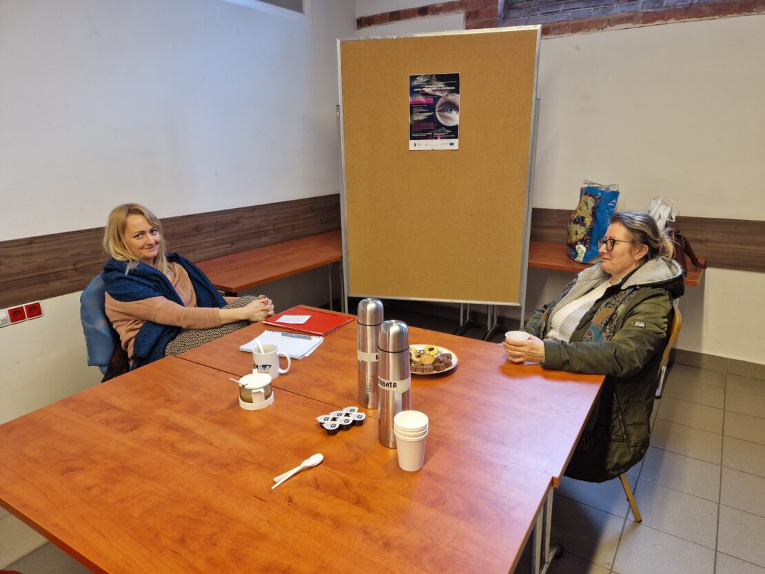 Stół przy nim siedzą dwie kobiety rozmawiające ze sobą. Na stole kubki z piciem i ciastka.