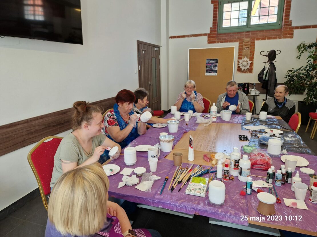 Grupa uczestników siedzi przy stole na którym jest wiele materiałów. Wszyscy malują na doniczkach
