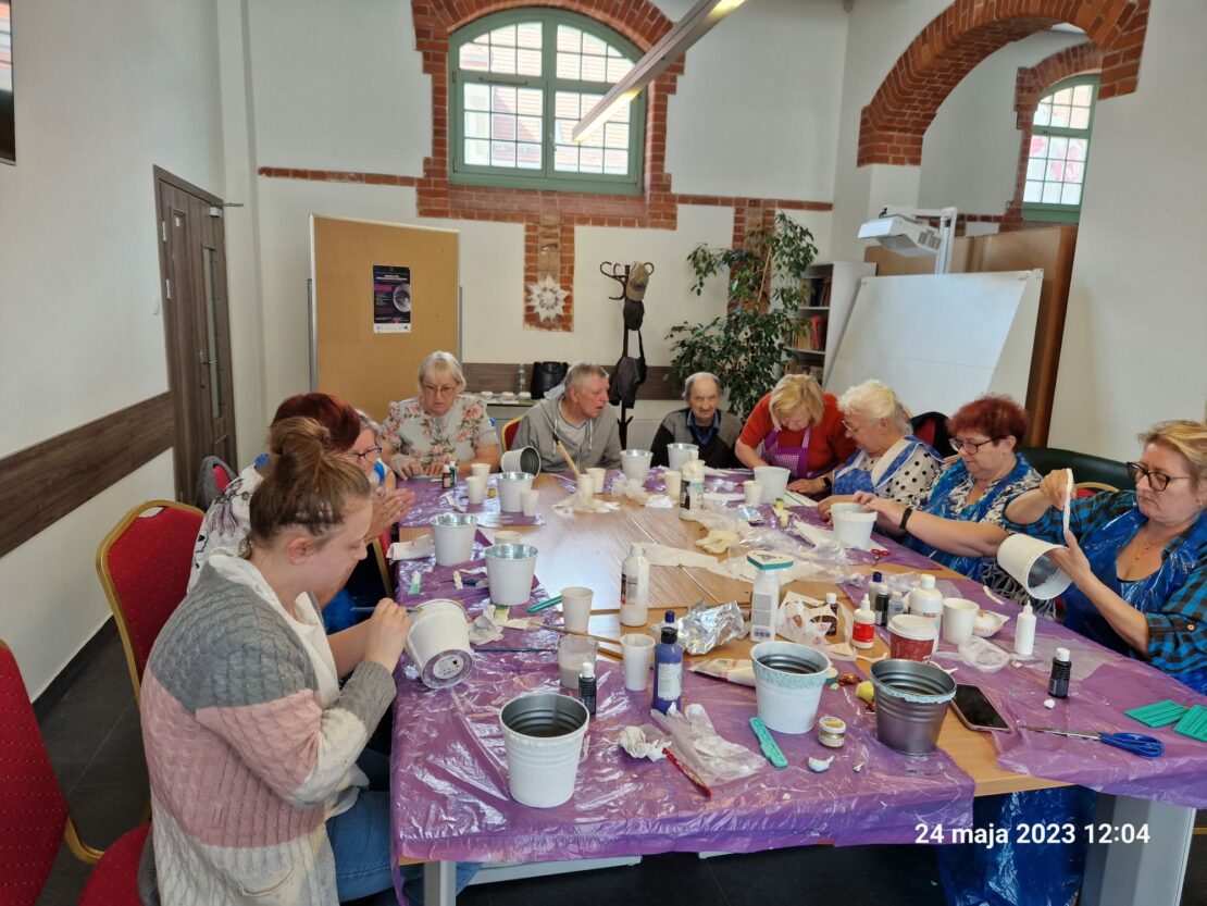 Stół na którym mamy wiele materiałów artystycznych, doniczki taśmy itp. dookoła siedzą uczestnicy szkolenia i malują na doniczkach