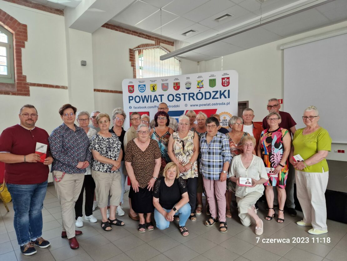 Grupowe zdjęcie wszystkich uczestników spotkania. Z tyłu baner informacyjny z napisem Powiat Ostródzki.