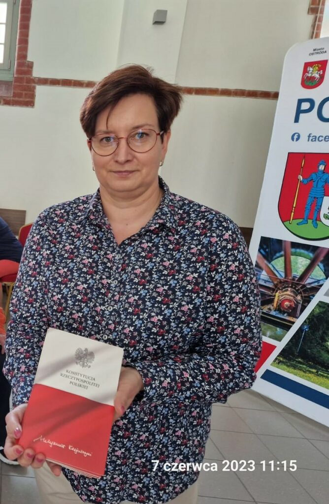 Pani Poseł trzyma w rękach Konstytucję Rzeczypospolitej Polskiej. Leki uśmiech z tyłu znajomy baner