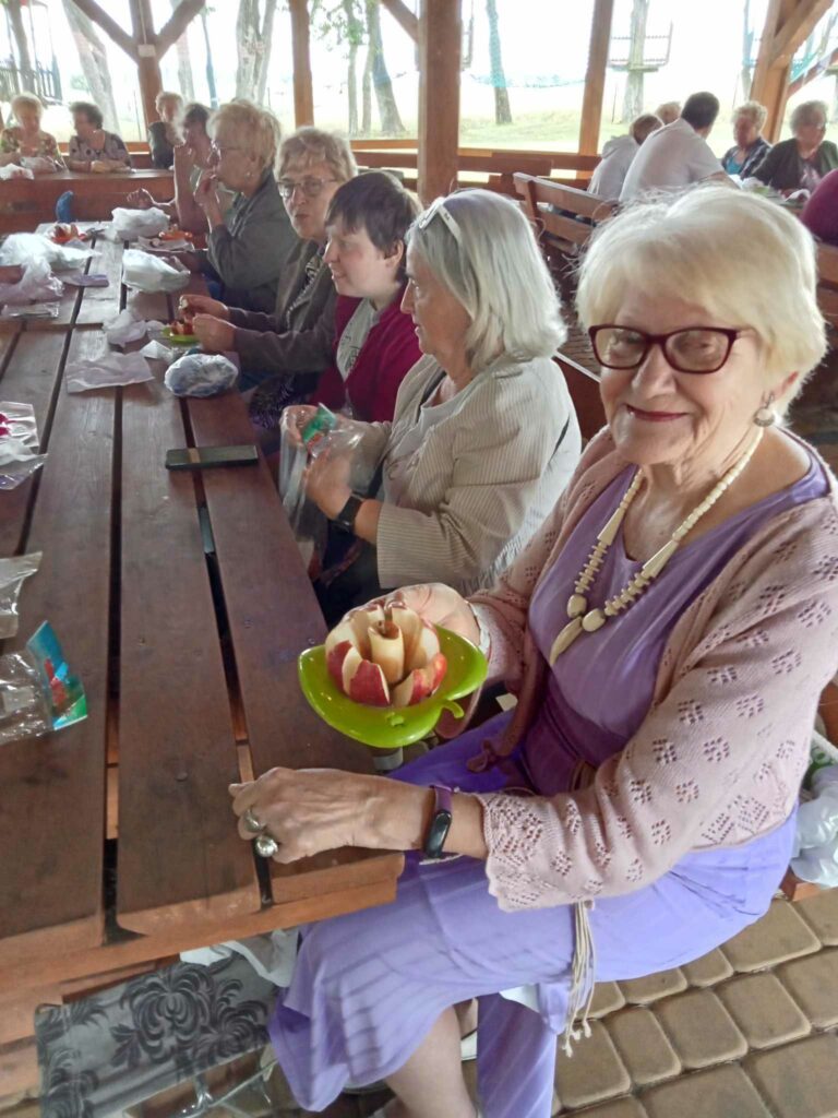 Grupa ludzi siedzi przy drewnianym stole w altance. Na pierwszym planie uśmiechnięta kobieta w ręku prezentuje  stworzonego kwiat z owoców