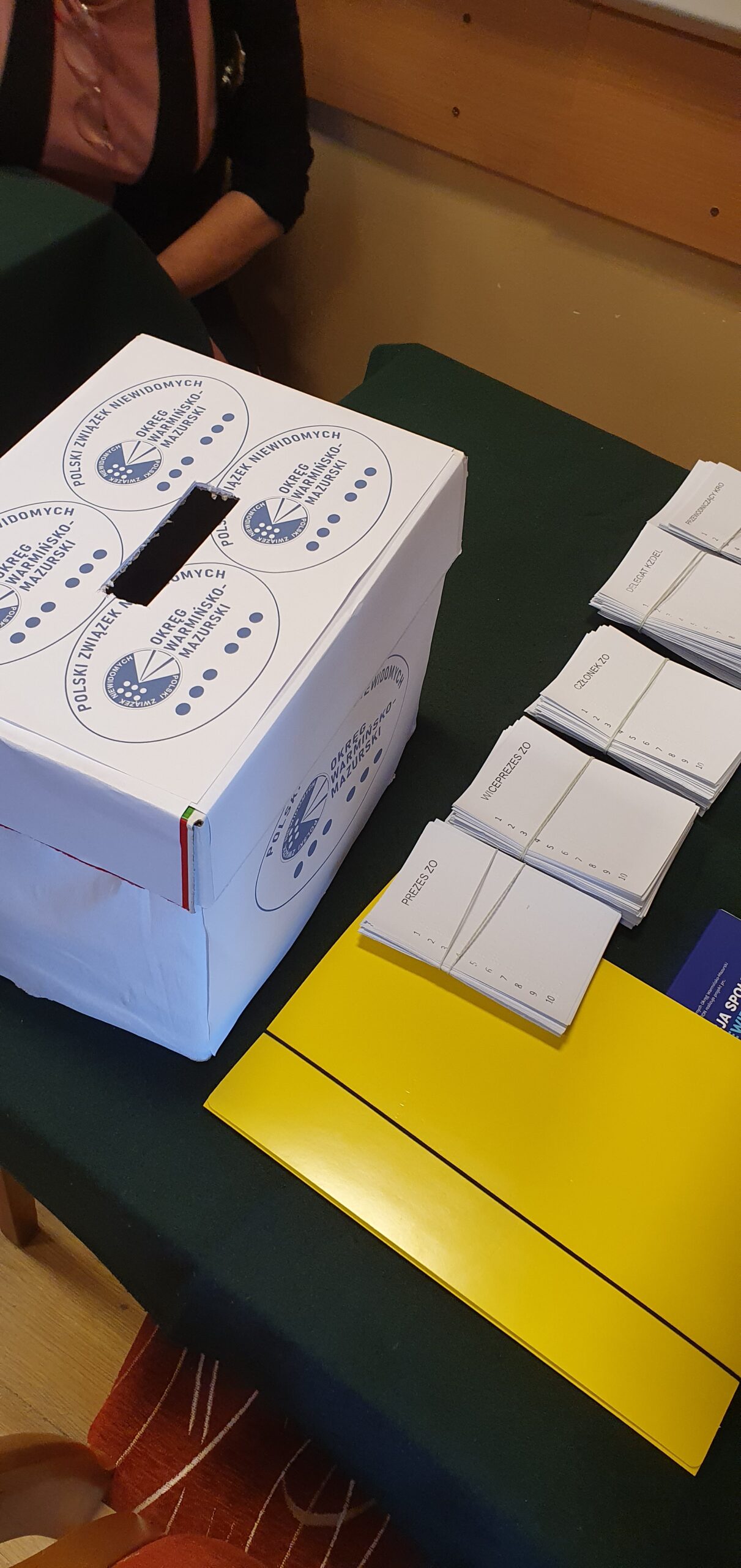Urna wyborcza a obok niej karty do głosowania w piśmie Braille'a oraz czarno druku