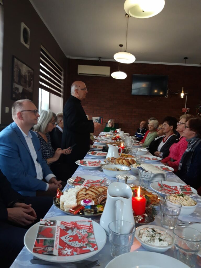 Sala restauracyjna. Ustawiono kilka stołów połączonych w 1. Jest on zasłany białym obrusem na nim talerze oraz dania. Przy nim siedzą ludzie 