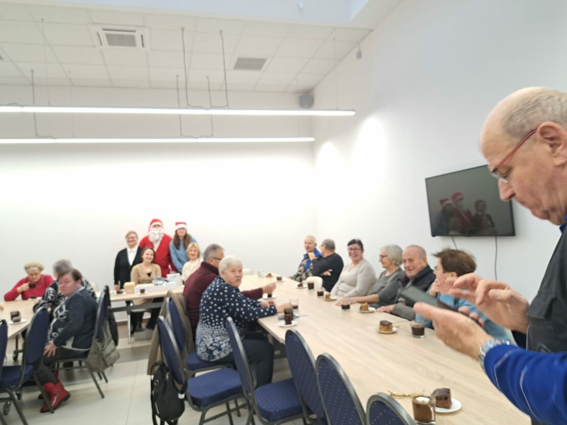 Sala konferencyjna, w środku ustawione stoły w literę "U"
Przy stołach siedzą ludzie  piją i jedzą ciasto.