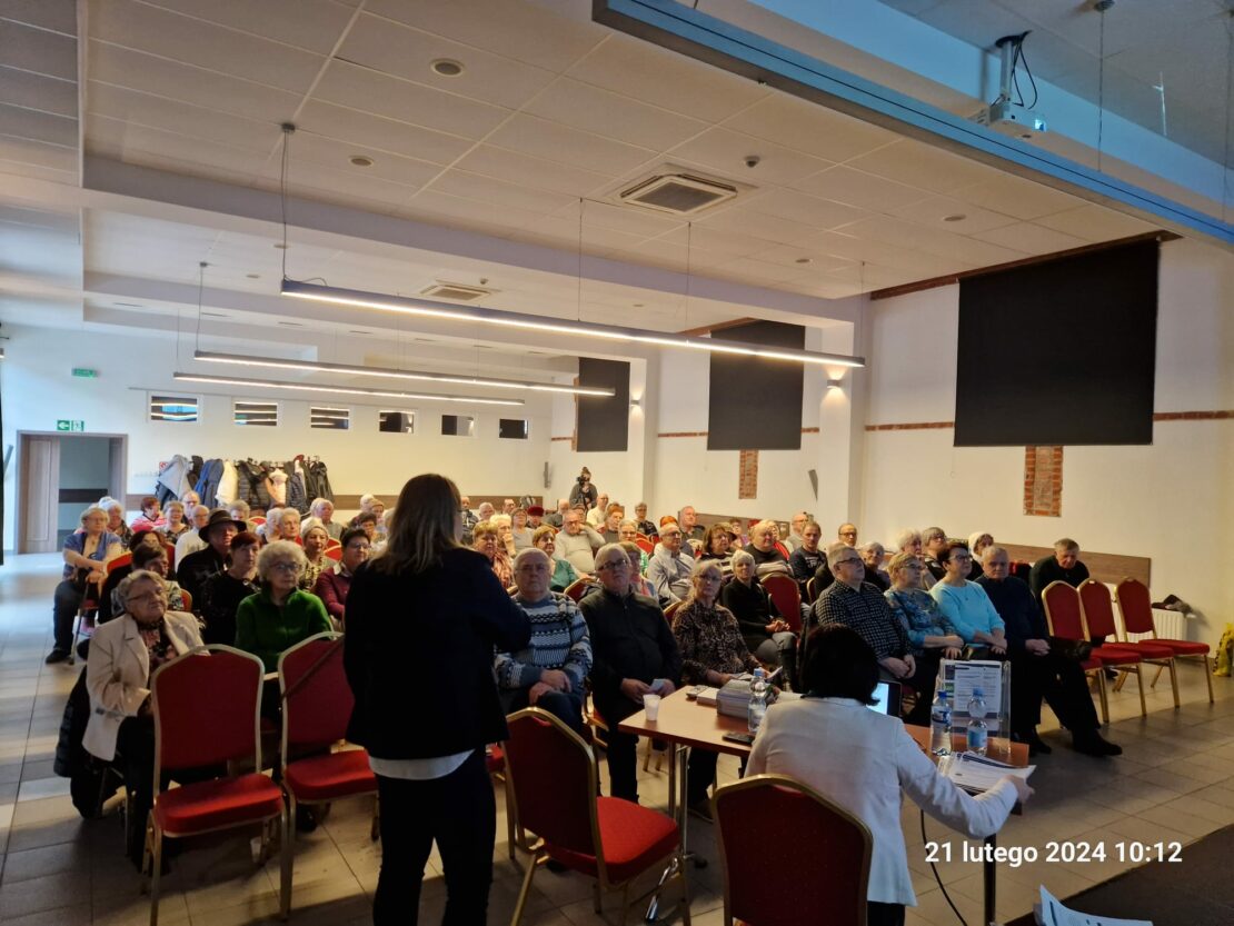 Ujęcie zza sceny na tłum uczestników siedzących i słuchających co Pani ma do powiedzenia.